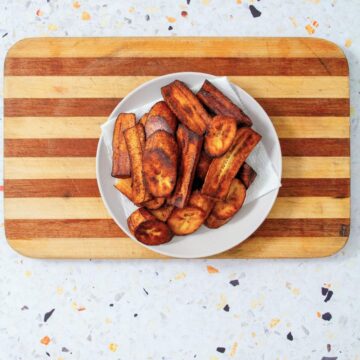A white bowl filled with caramelized fried sweet plantains rests on a striped wooden cutting board against a speckled terrazzo surface.