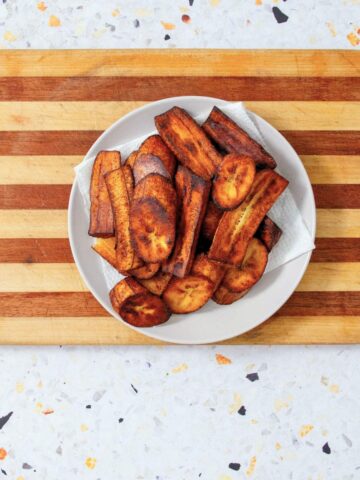 A white bowl filled with caramelized fried sweet plantains rests on a striped wooden cutting board against a speckled terrazzo surface.