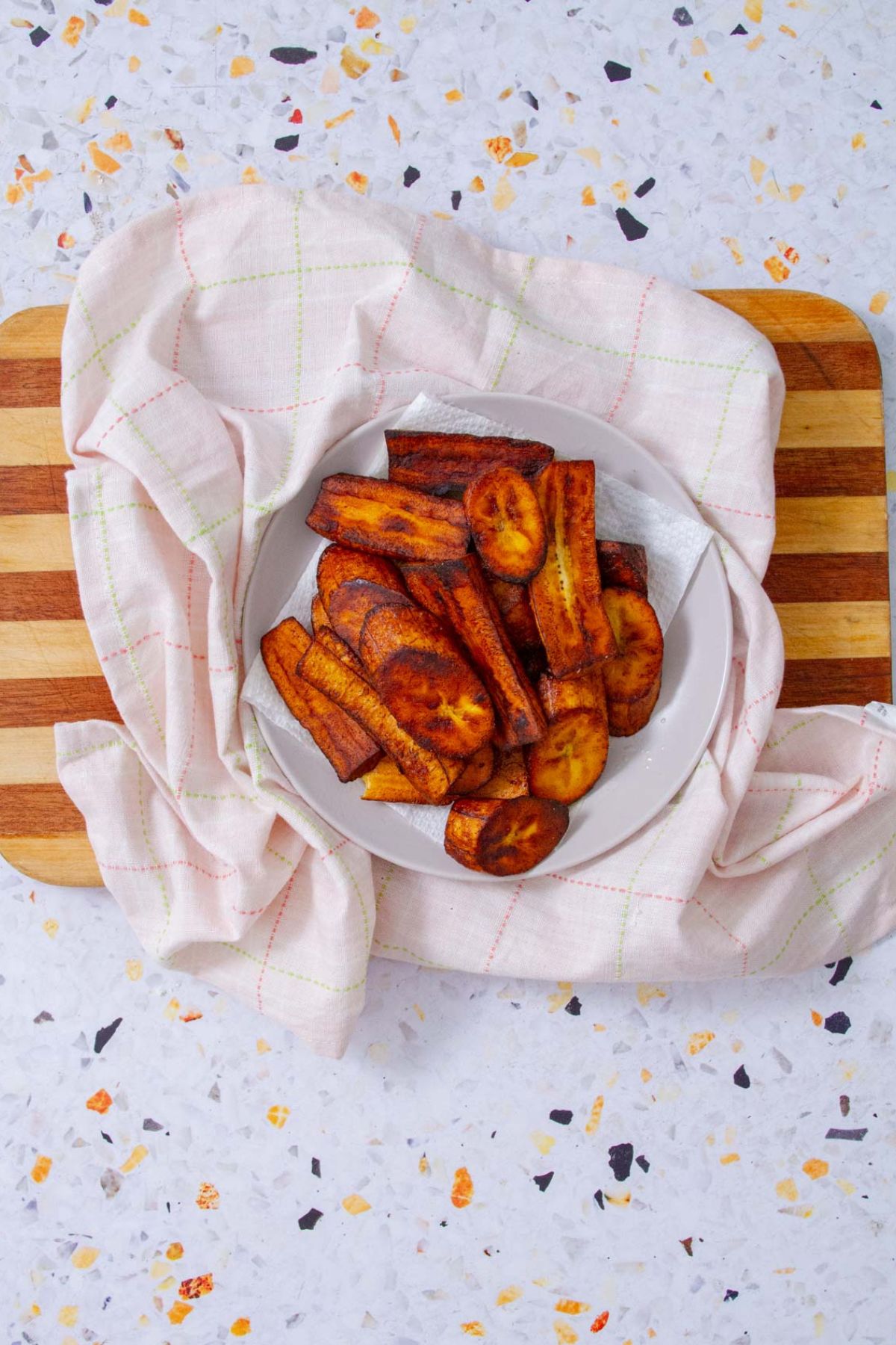Caramelized amarillos piled in a white bowl lined with paper towels, draped in a pink checkered kitchen towel on a striped wooden cutting board, viewed from overhead.