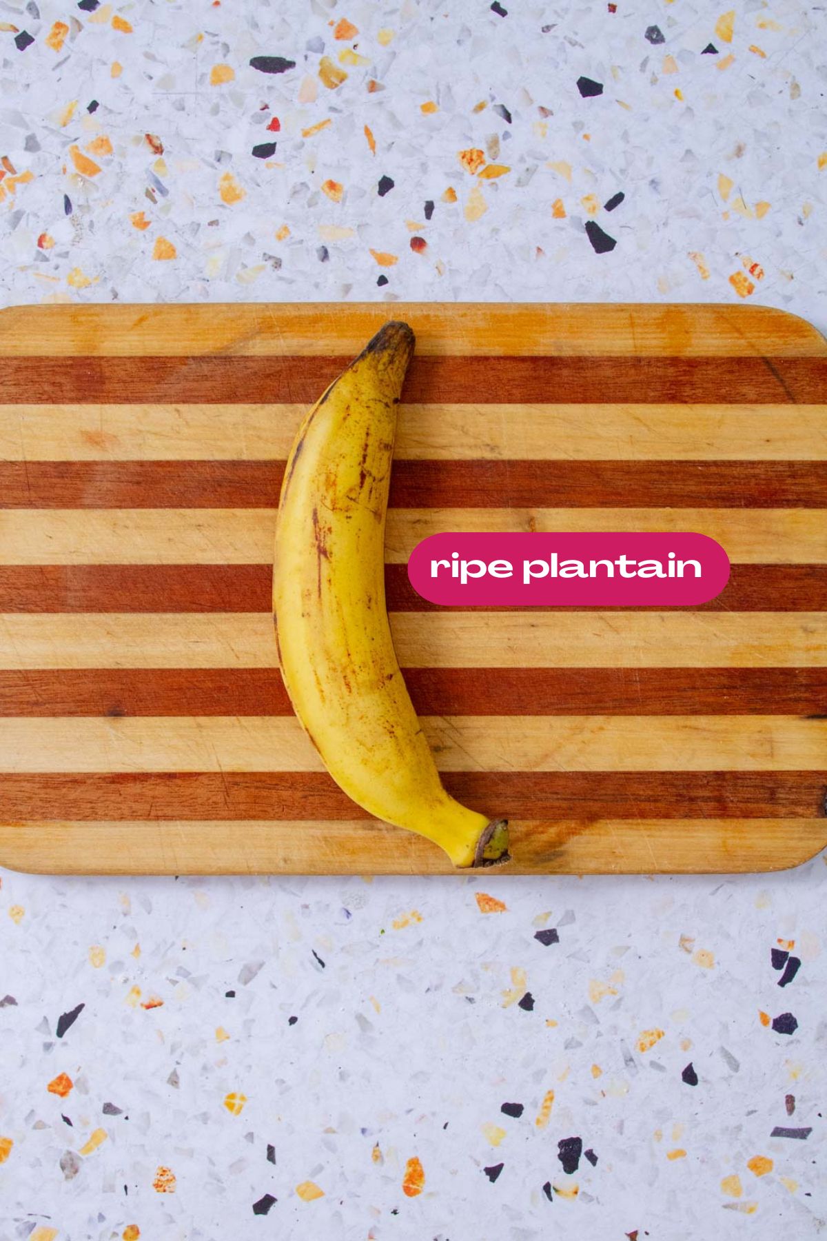 A ripe yellow plantain with brown speckles resting on a striped wooden cutting board against a light terrazzo surface, labeled "ripe plantain."