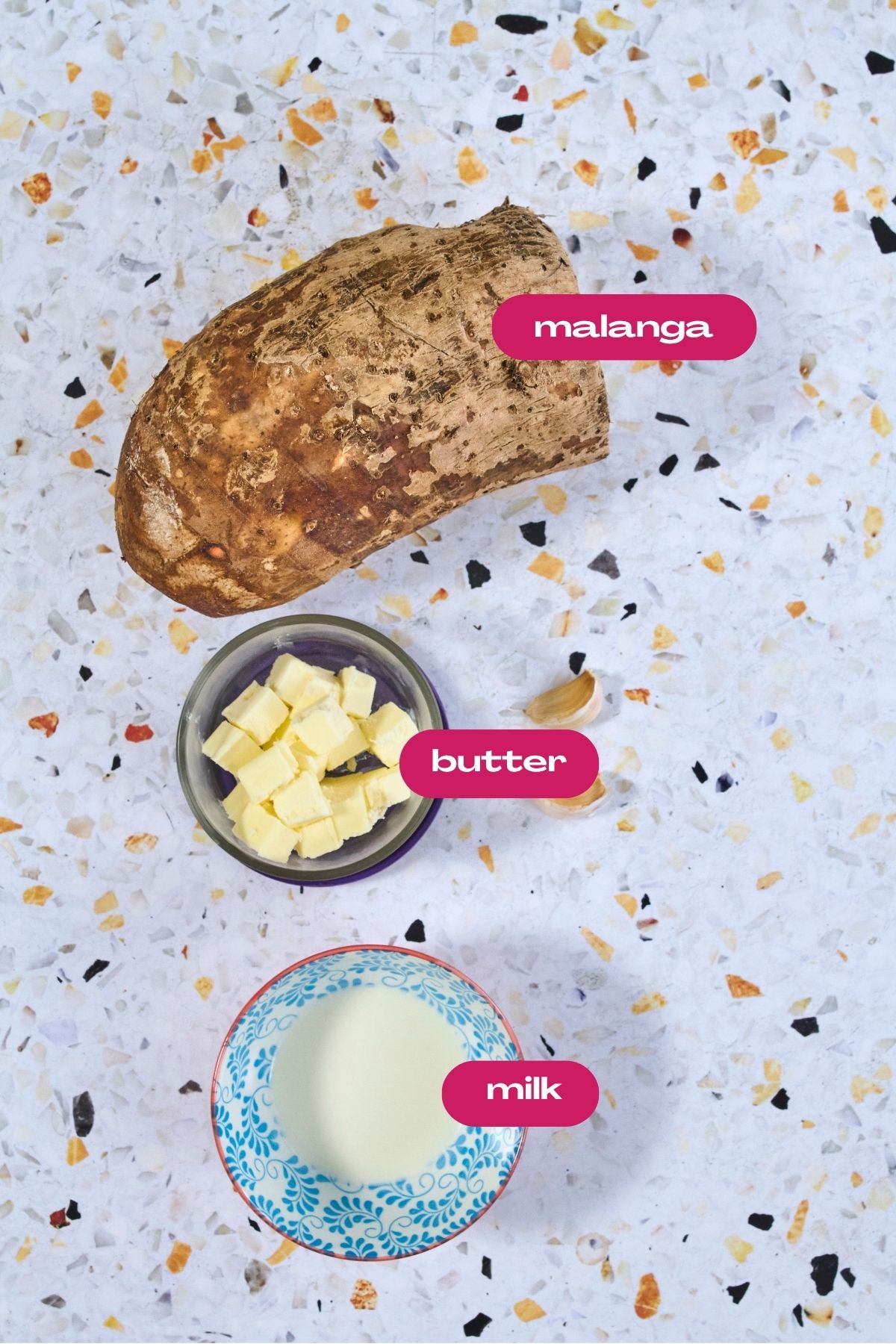 Ingredients for mashed malanga: whole malanga corm, cubed butter in a bowl, and milk in a blue floral bowl on a terrazzo surface.