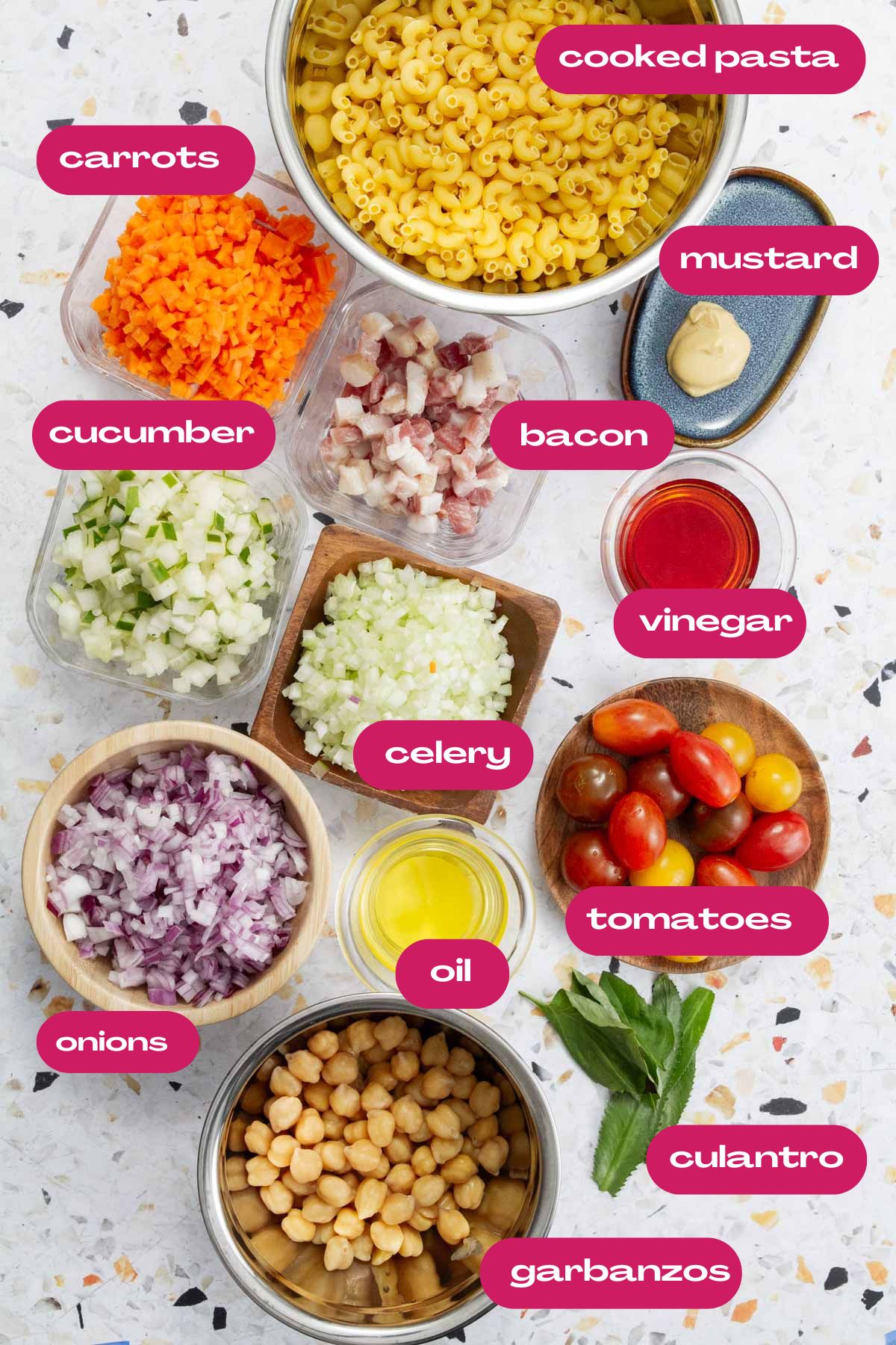 All ingredients for pasta salad laid out, including cooked macaroni, diced carrots, cucumber, celery, red onions, cherry tomatoes, garbanzos, bacon, olive oil, vinegar, mustard, and fresh cilantro.