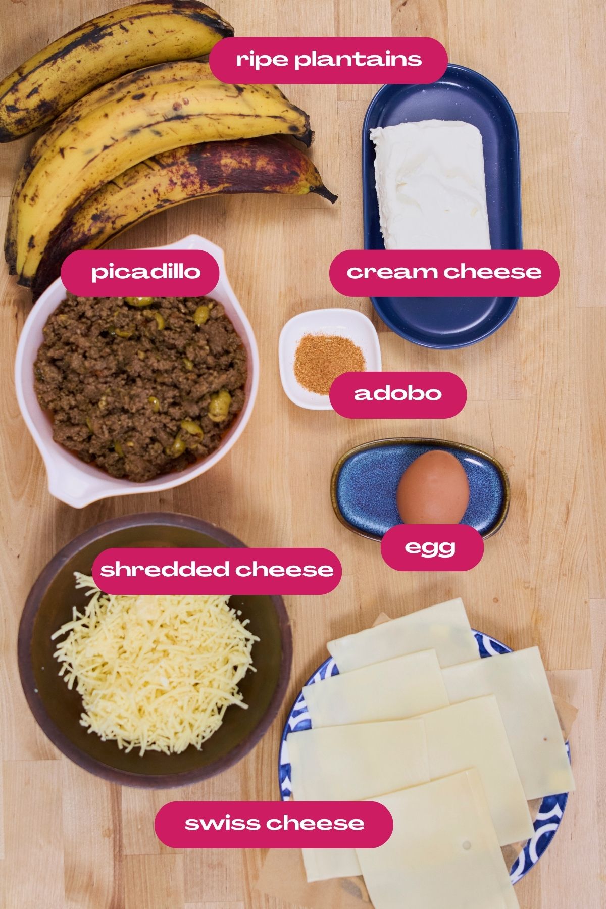 Labeled ingredients for pastelón arranged on a wood surface: ripe plantains, picadillo, cream cheese, adobo seasoning, egg, shredded cheese, and sliced Swiss cheese.