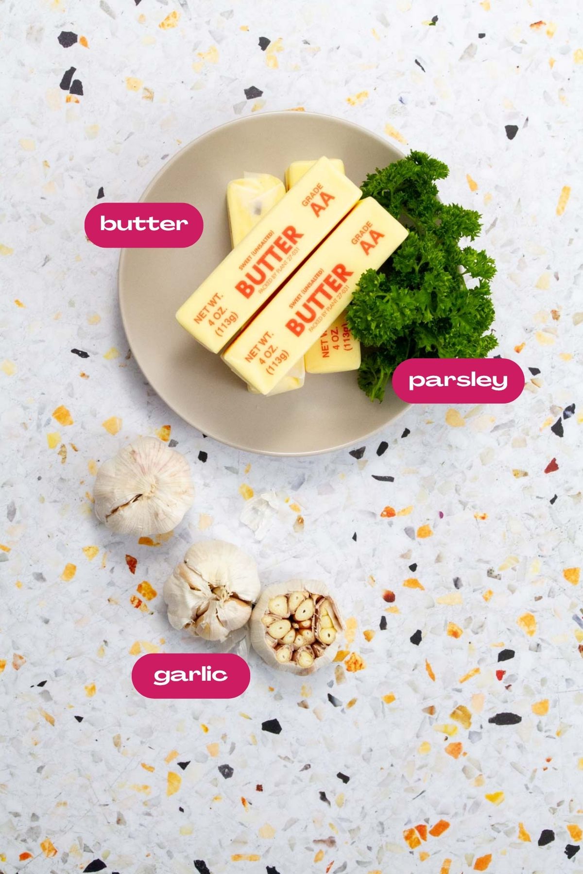 Ingredients for roasted garlic and herb compound butter, including butter sticks, fresh parsley, and garlic heads on a terrazzo surface.