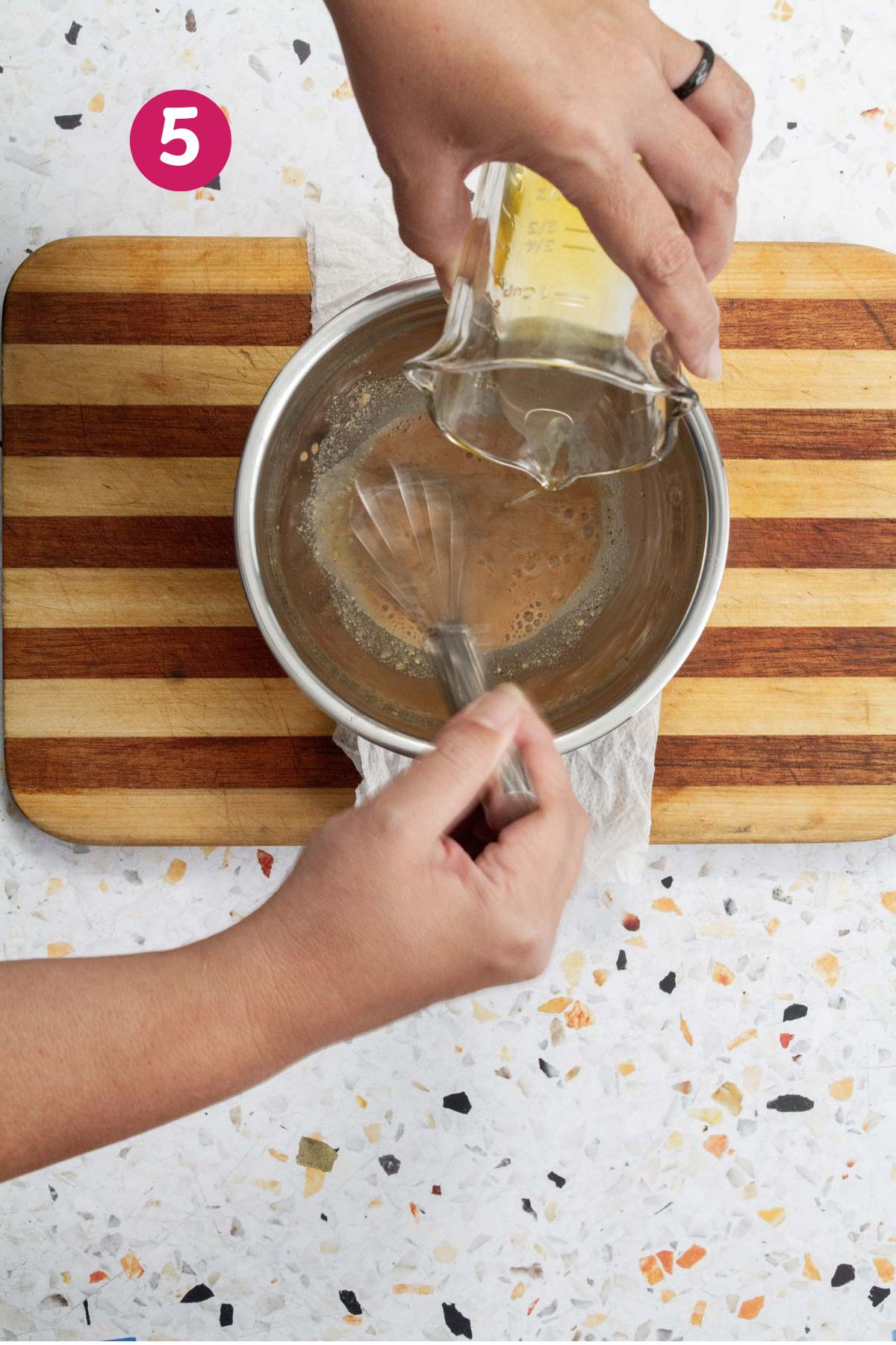 Whisking olive oil into the vinaigrette dressing in a stainless steel bowl on a wooden cutting board.