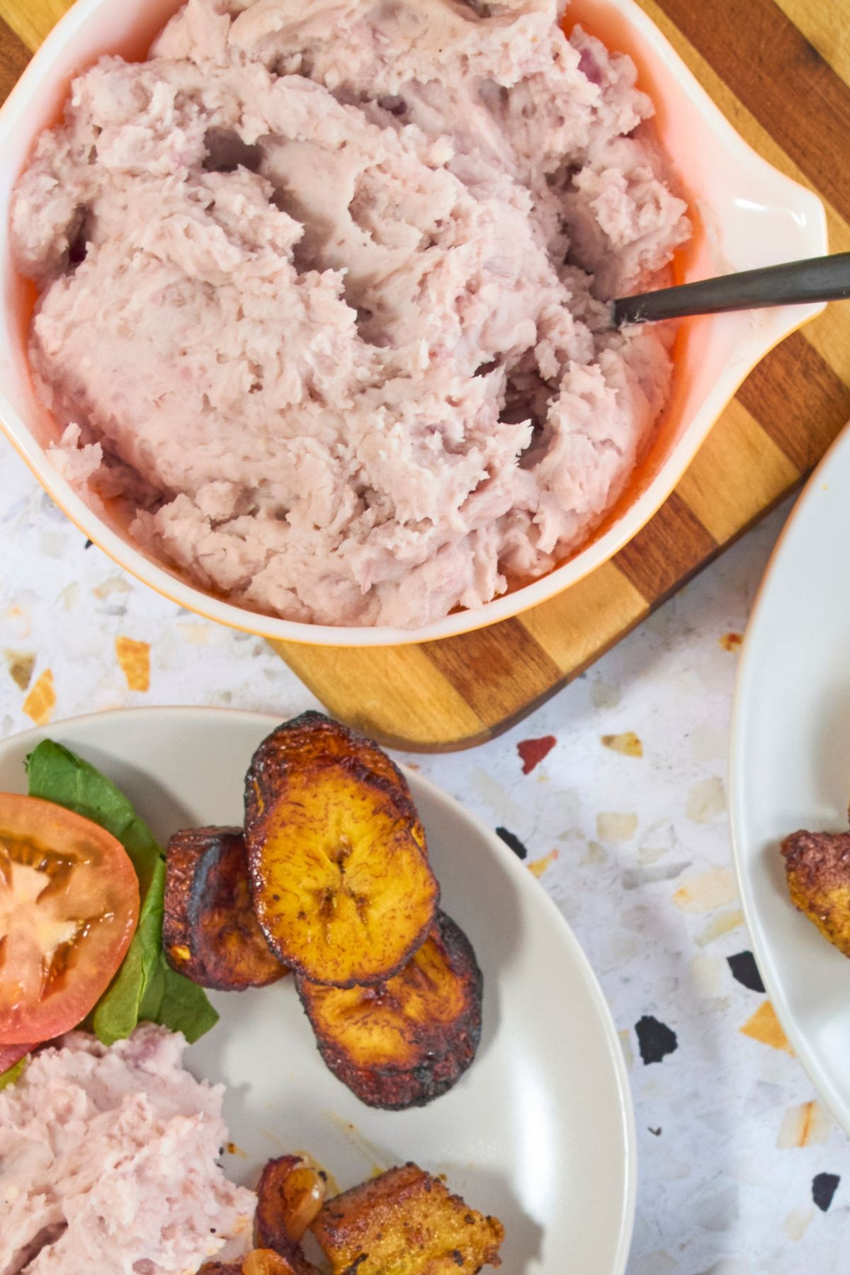 Close up of mashed malanga in a vintage orange Pyrex dish with a serving spoon, next to a plate with maduros and tomato slices.