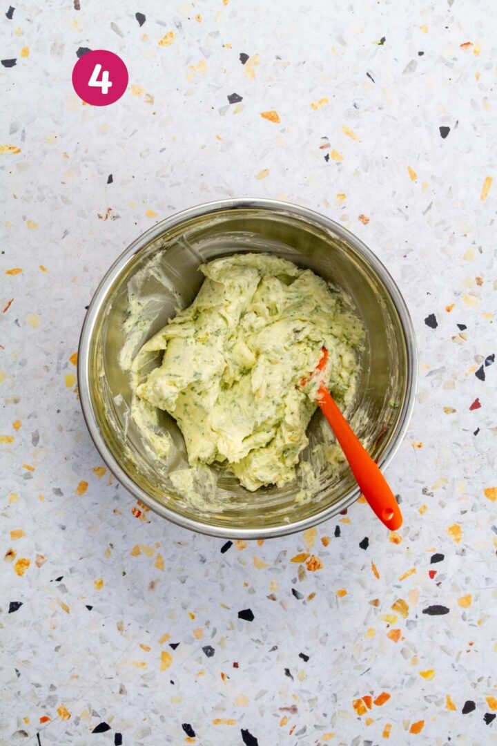 Fully mixed roasted garlic and herb compound butter in a stainless steel bowl with an orange spatula.
