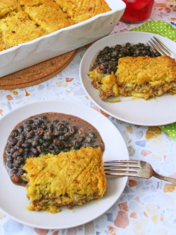 (terrazzo surface, two plates + baking dish) Two plates of pastelón served with black beans on a terrazzo surface, with the white baking dish and colorful flowers in the background.