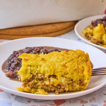 Close-up of a slice of pastelón on a white plate showing the mashed plantain crust and seasoned picadillo filling, served alongside black beans.
