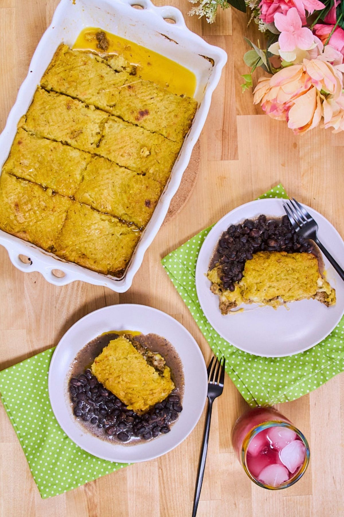 (overhead, flowers, wood surface) Overhead view of pastelón in a white baking dish scored into portions, with two plated servings on green polka dot napkins and pink flowers in the corner.