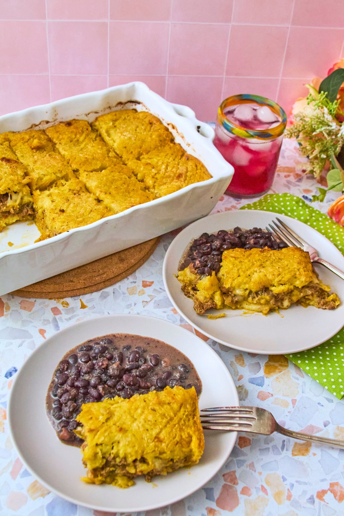 (terrazzo, pink tile background, vertical) Pastelón in a white baking dish on a terrazzo surface with two plated servings alongside black beans, a colorful glass of iced drink, and fresh flowers.