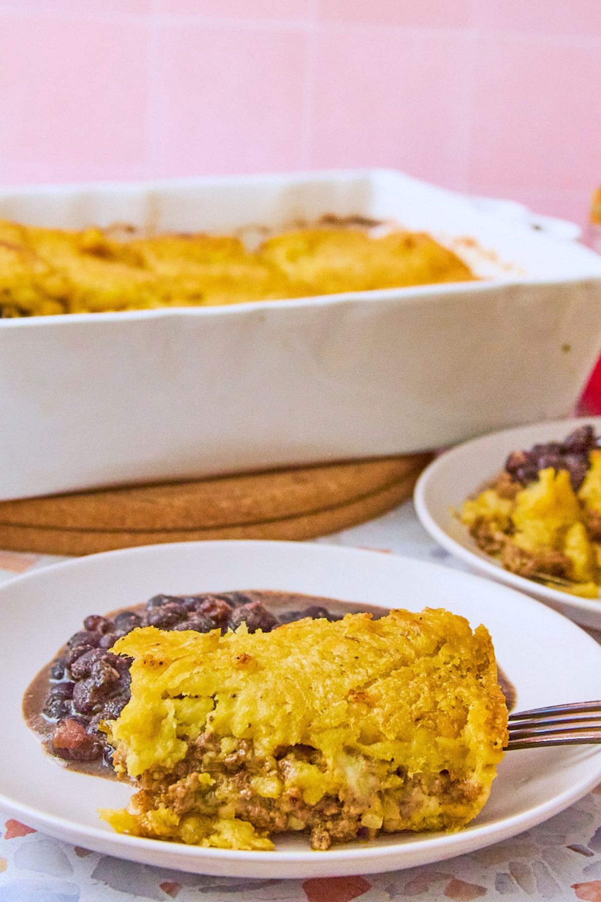 (pink tile, close-up slice, vertical) A slice of pastelón on a white plate showing the golden mashed plantain crust and picadillo filling, with the white baking dish blurred in the background.