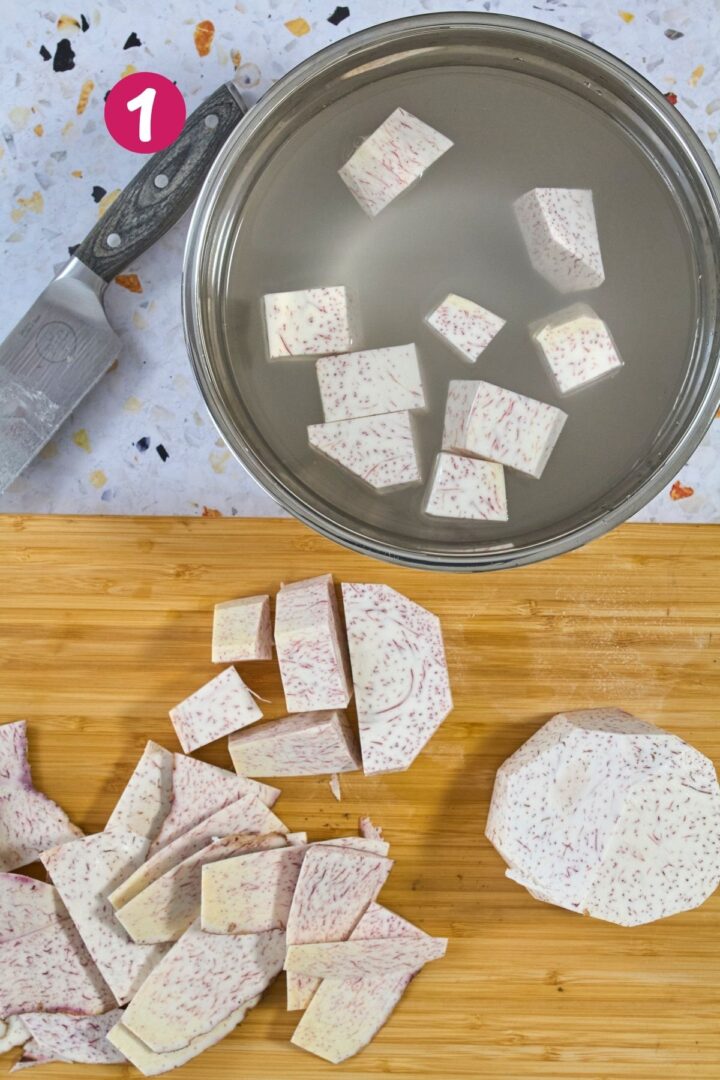 Peeled and cubed raw malanga on a bamboo cutting board next to a stainless steel pot of water.