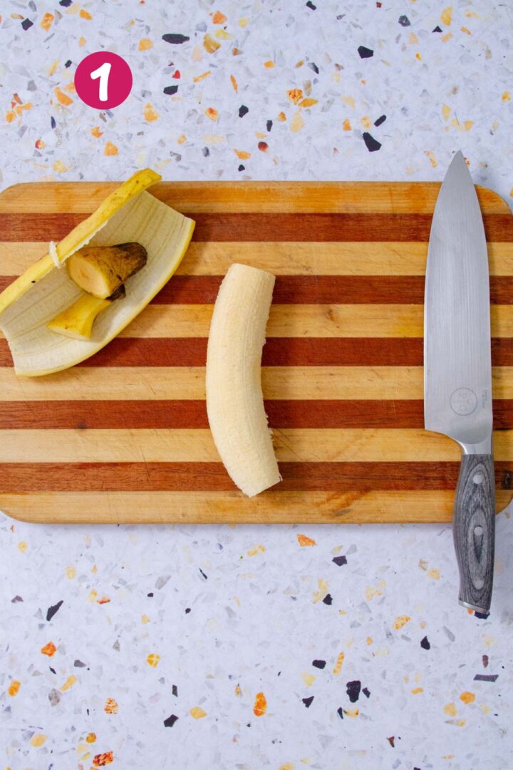 A whole peeled ripe plantain on a striped wooden cutting board next to its yellow skin, ready to be sliced, with a chef's knife resting alongside on a speckled terrazzo surface.