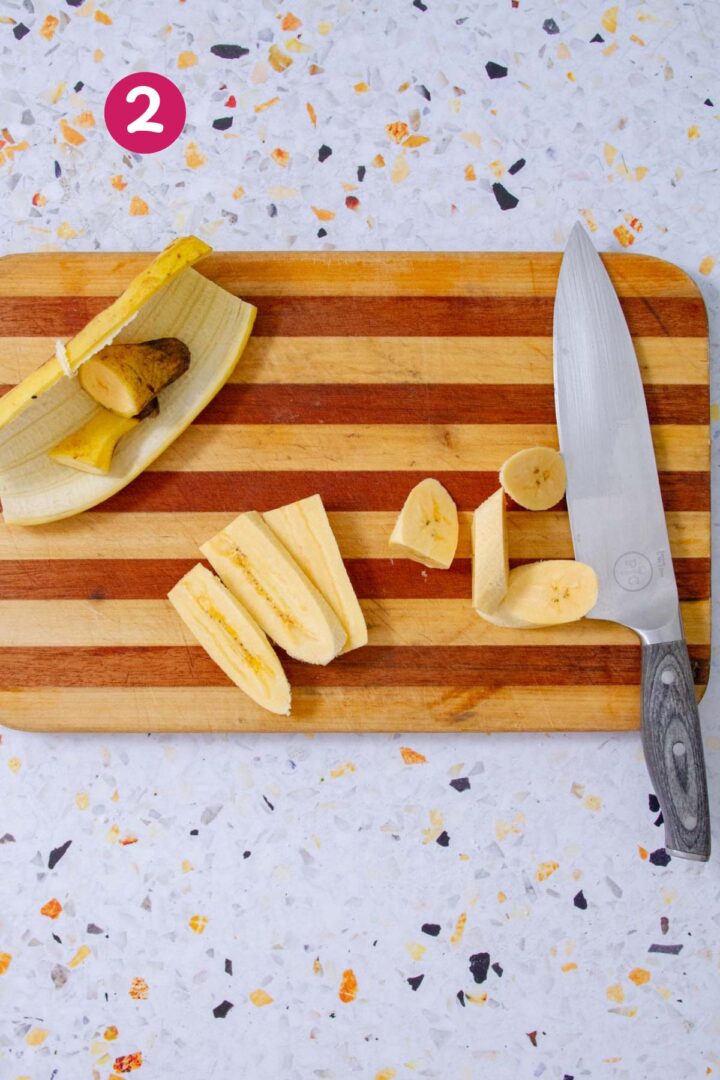 A peeled ripe plantain sliced into diagonal pieces on a striped wooden cutting board, with the removed peel set aside and a chef's knife nearby, on a speckled terrazzo surface.