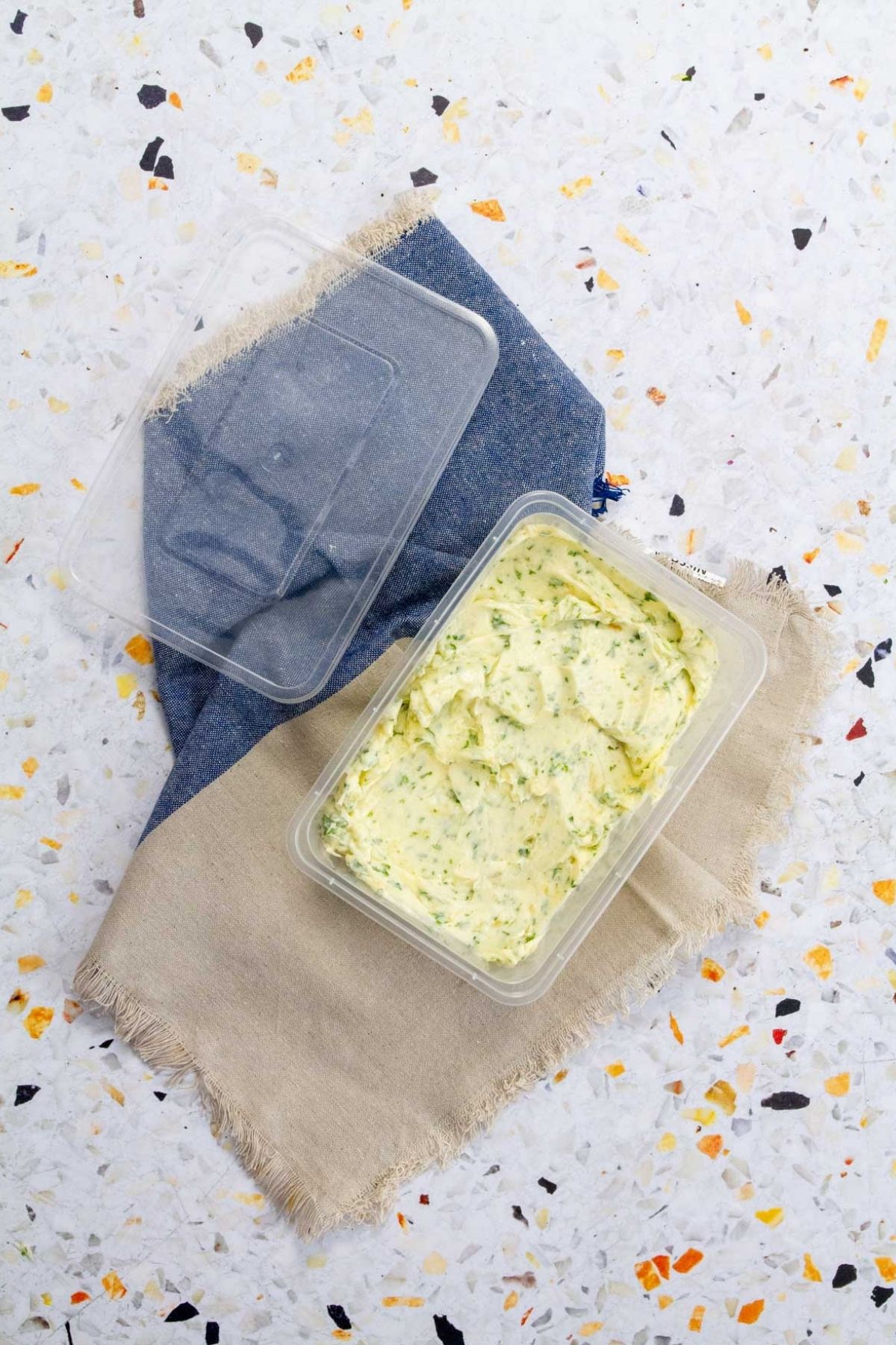 Roasted garlic and herb compound butter in an open, clear storage container on a blue and tan linen.