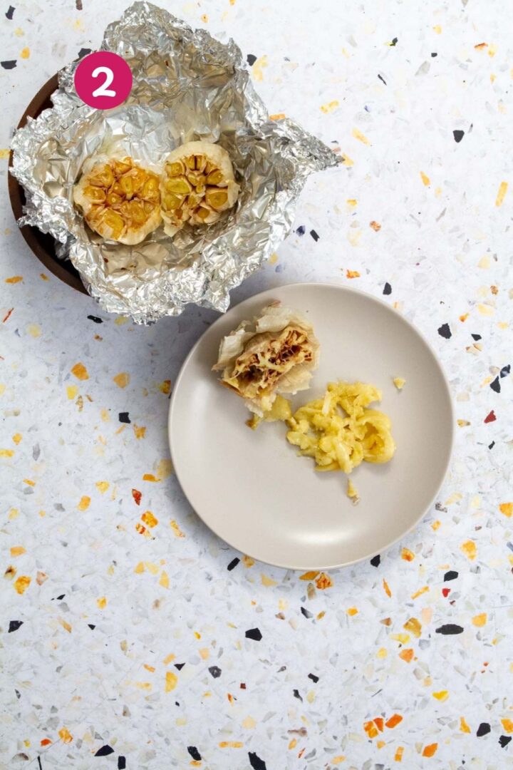 Roasted garlic heads in open foil next to squeezed and mashed roasted garlic cloves on a small plate.