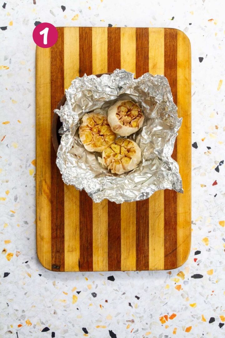 Three roasted garlic heads in opened aluminum foil on a wooden cutting board.