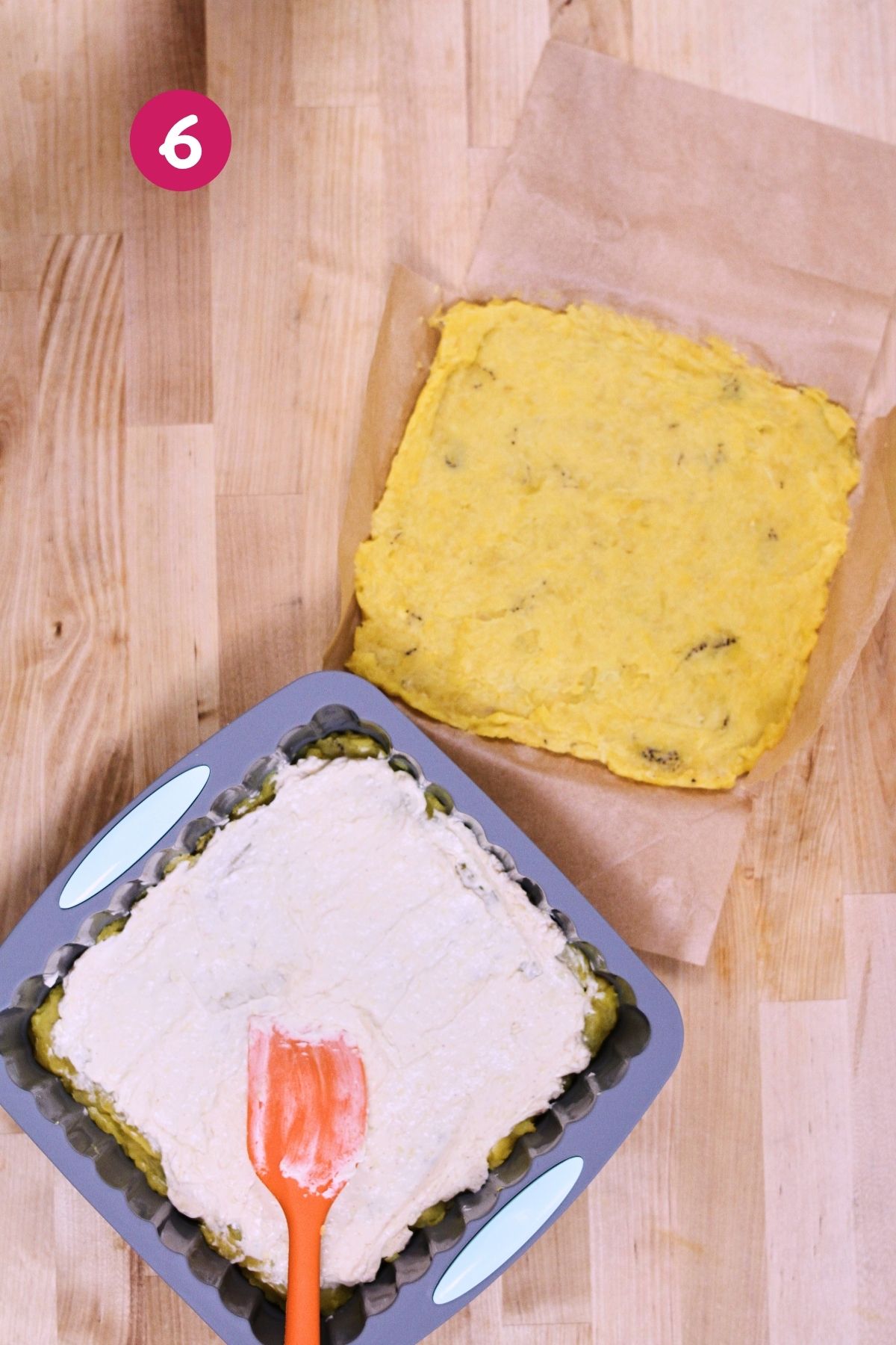 (Step 6) Cream cheese mixture being spread over the bottom layer of mashed plantains in a parchment-lined baking pan, with the top plantain layer resting on parchment paper alongside.