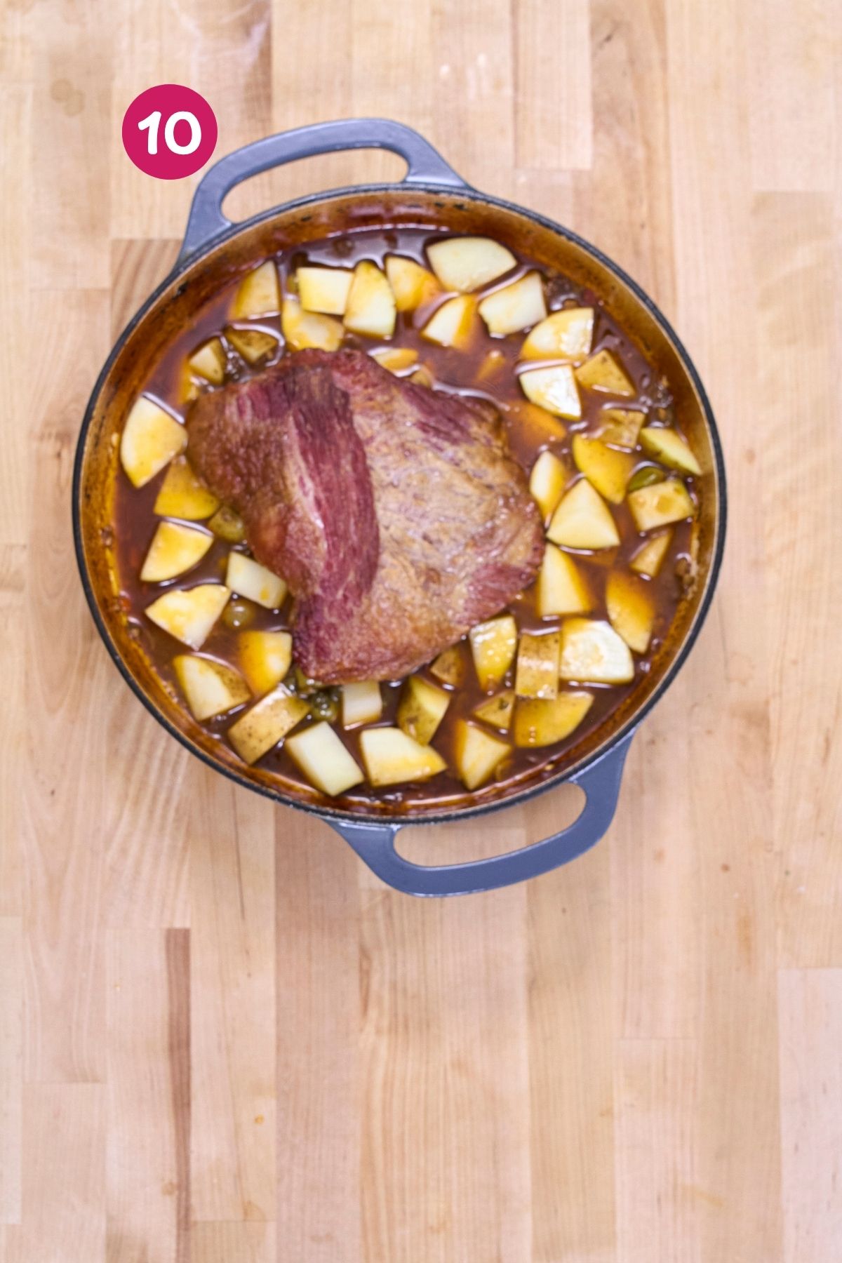 Browned corned beef brisket surrounded by cubed potatoes in seasoned broth in a Dutch oven.
