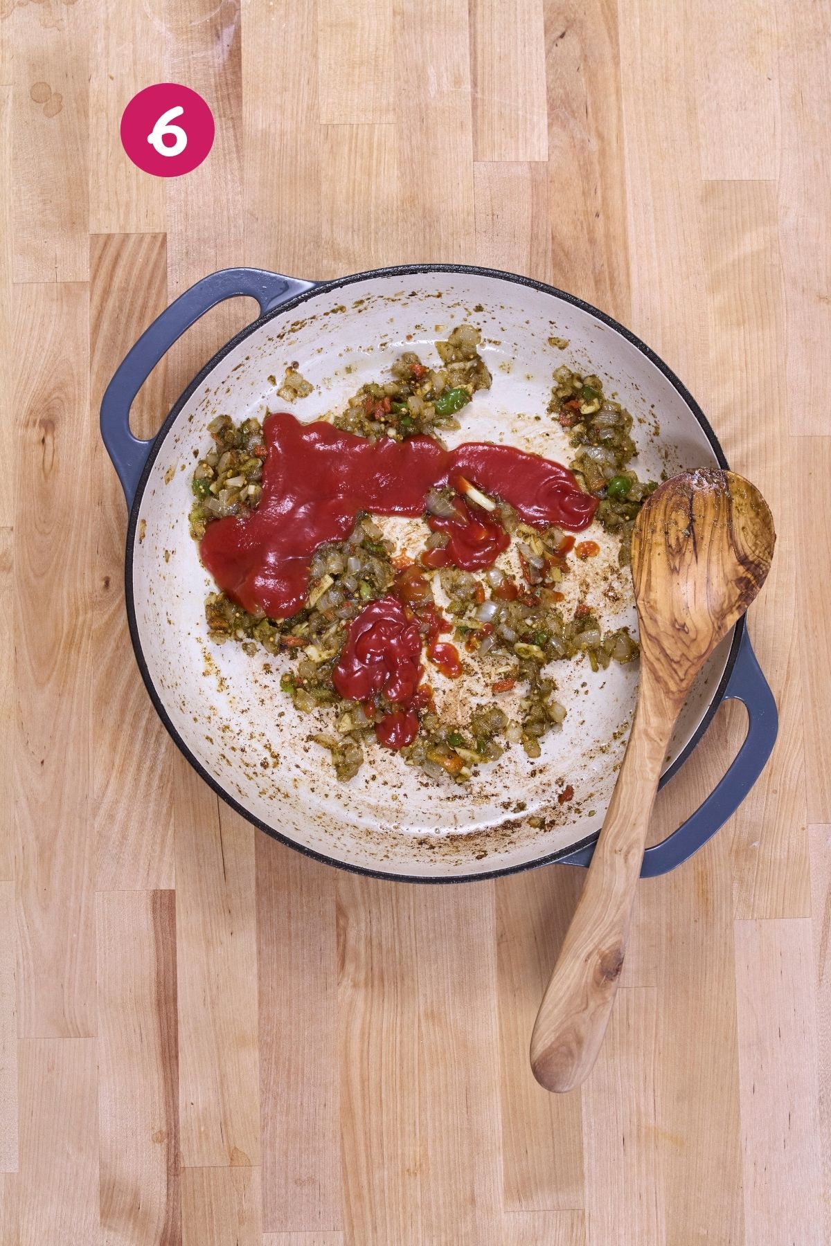 Tomato sauce and sofrito are added to sautéed onions, ají dulce, and garlic in an enameled pan with a wooden spoon.
