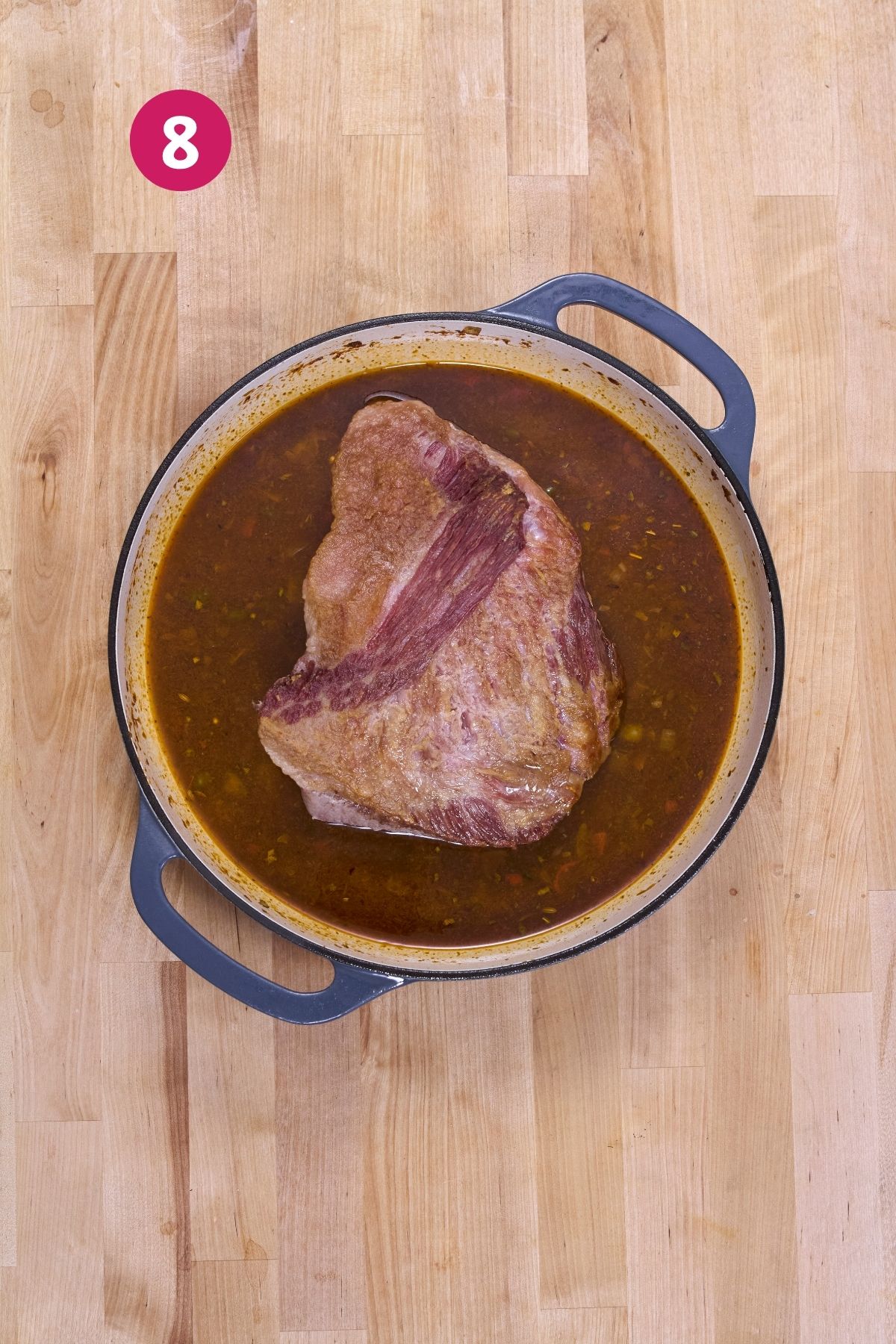 Browned corned beef brisket simmering in a deep reddish-brown broth in an enameled Dutch oven.