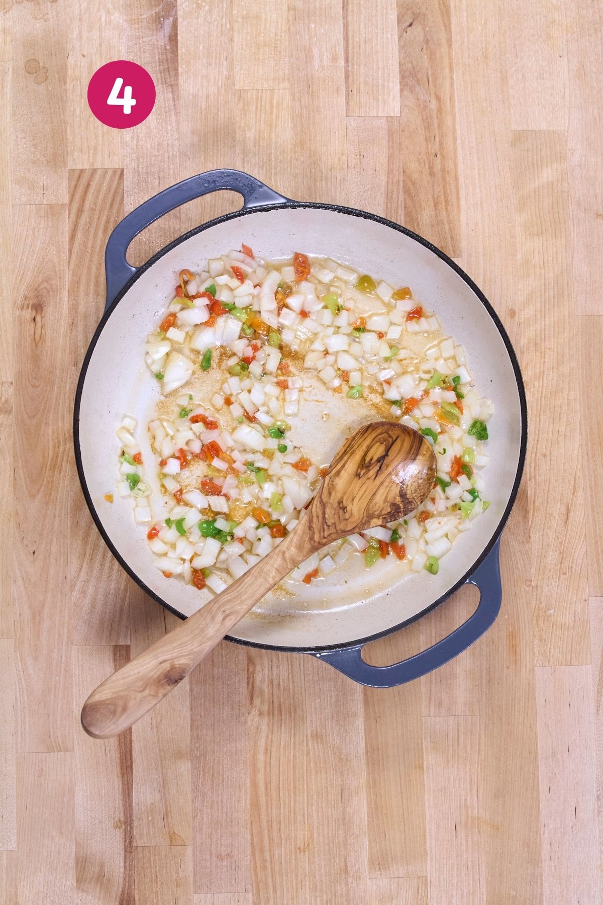 Diced onions and ají dulce peppers are sautéing in an enameled cast-iron pan with a wooden spoon.
