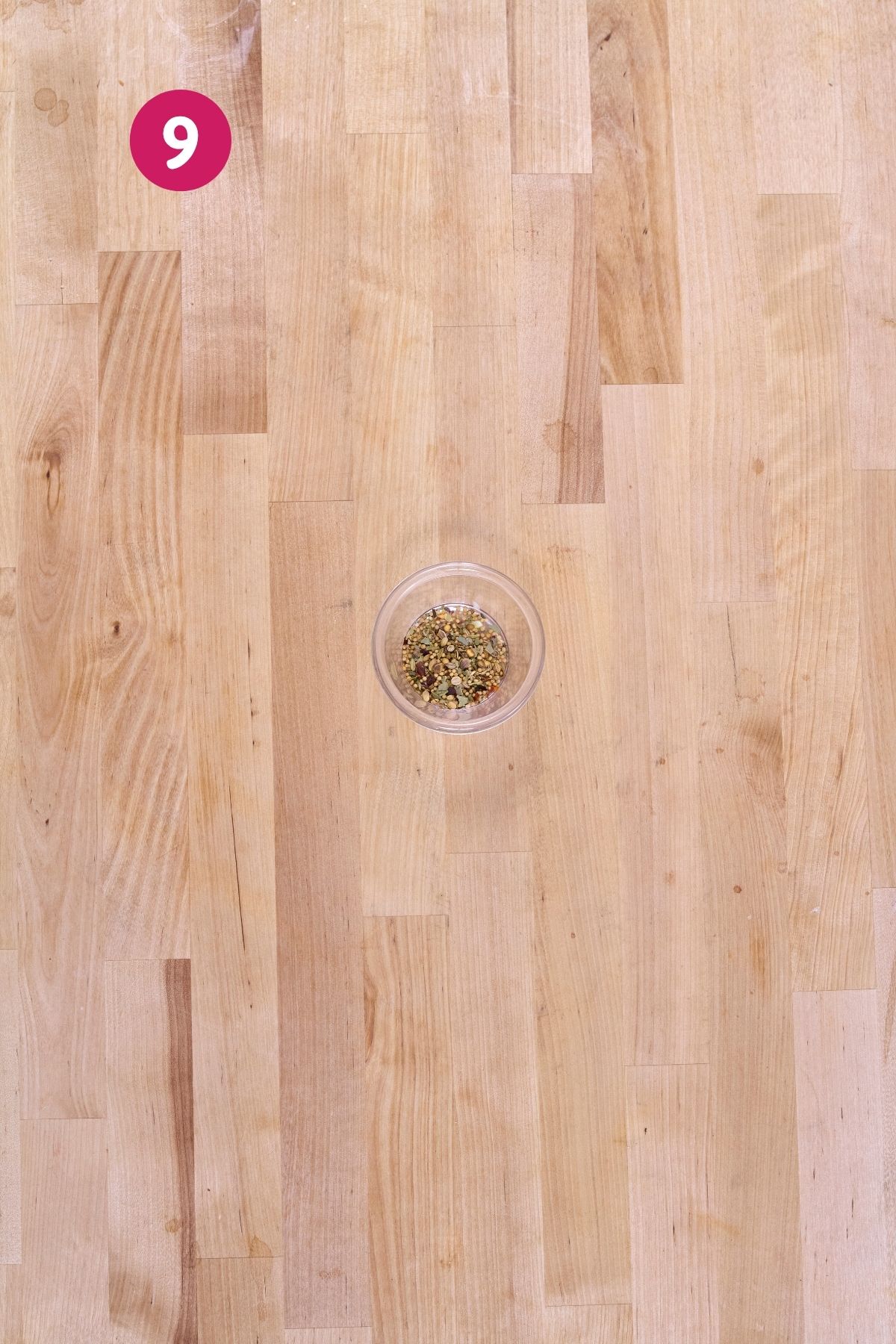 Small glass bowl of pickling spices with mustard seeds, coriander, bay, and red pepper flakes on a butcher block counter.