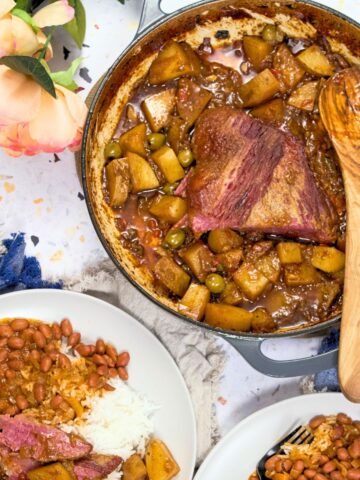 Puerto Rican corned beef brisket with potatoes and olives in a Dutch oven, served alongside plates of white rice and stewed beans.
