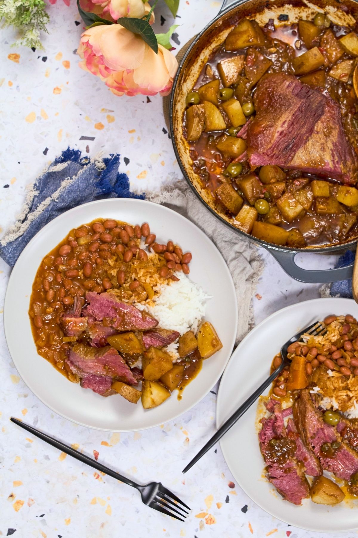 Overhead view of Puerto Rican corned beef brisket in a Dutch oven with potatoes, olives, and capers, served with rice and stewed beans.