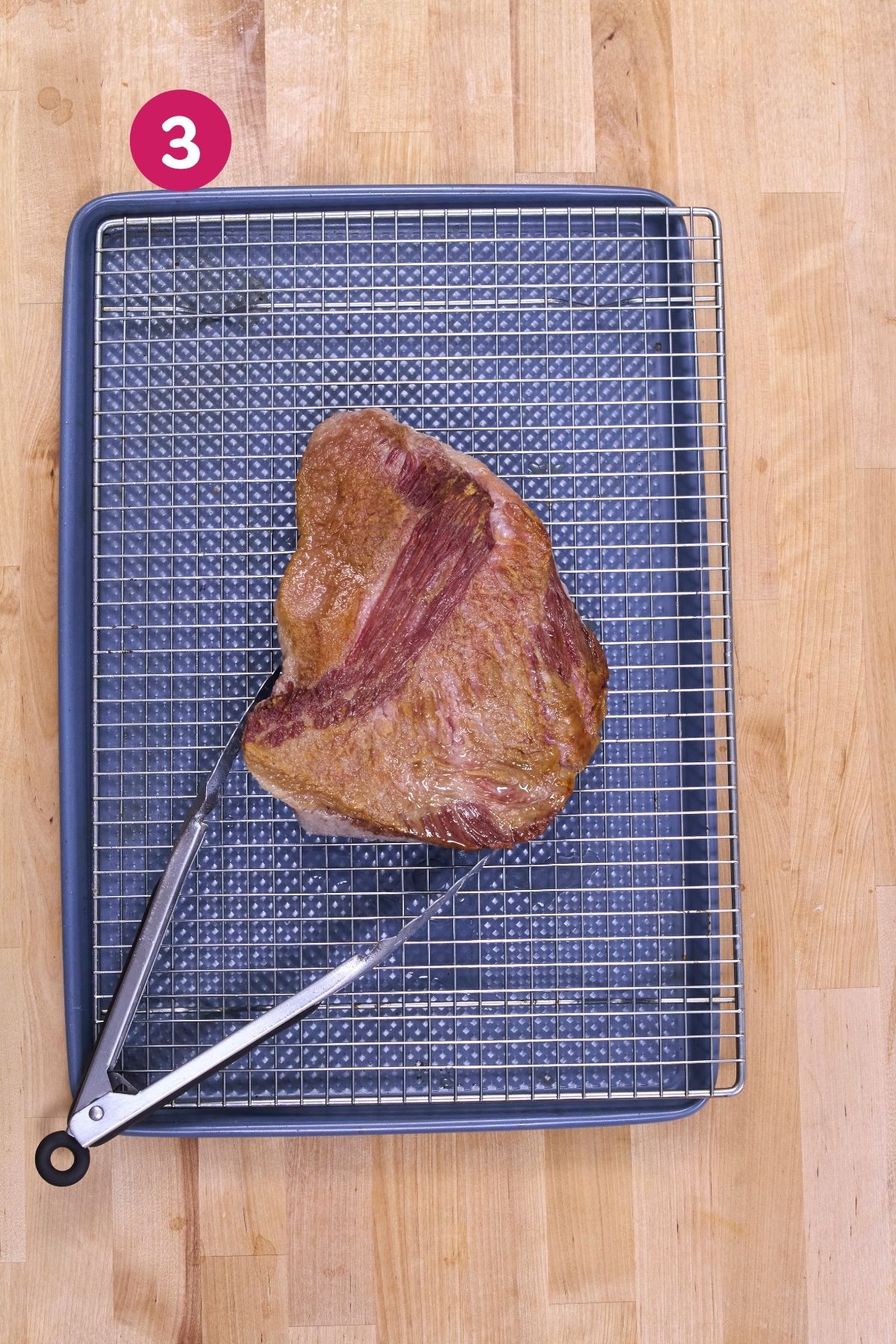 Browned corned beef brisket resting on a wire rack over a baking sheet, tongs nearby.