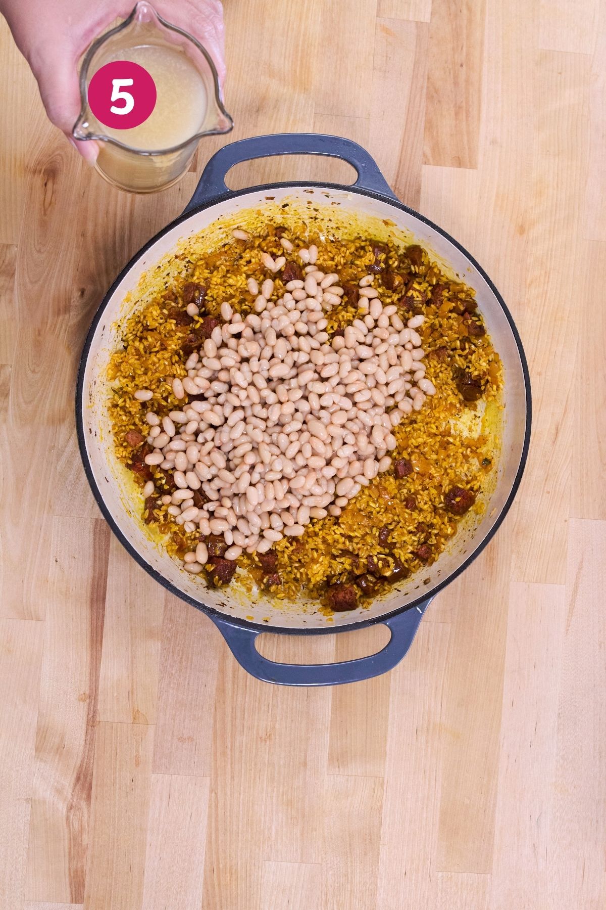 Pork broth being poured into a pot of rice, chorizo, and white beans for arroz con chorizo y habichuelas blancas.