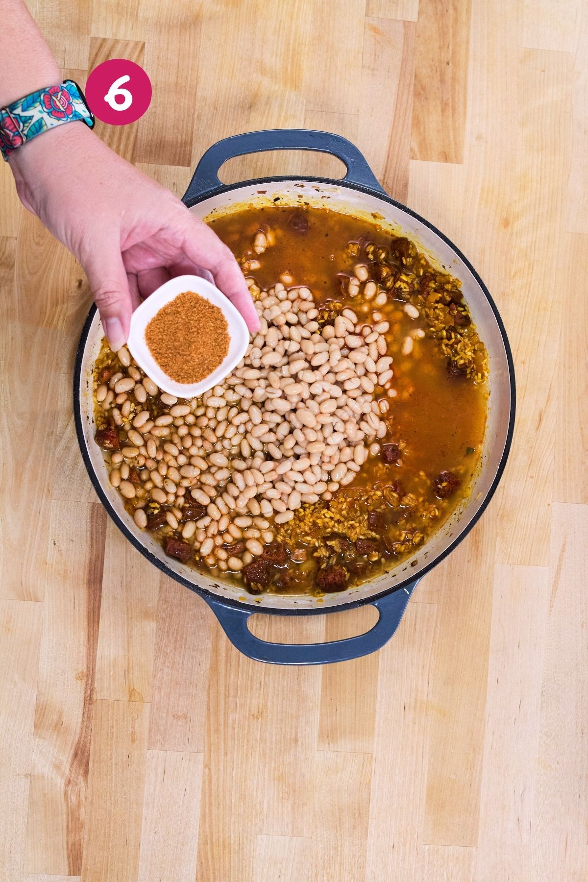 Adobo seasoning is being added to a pot of simmering white beans, rice, and chorizo.