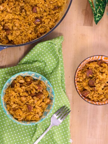 Two bowls of arroz con chorizo y habichuelas blancas served beside the Dutch oven, with forks and a green polka-dot napkin.